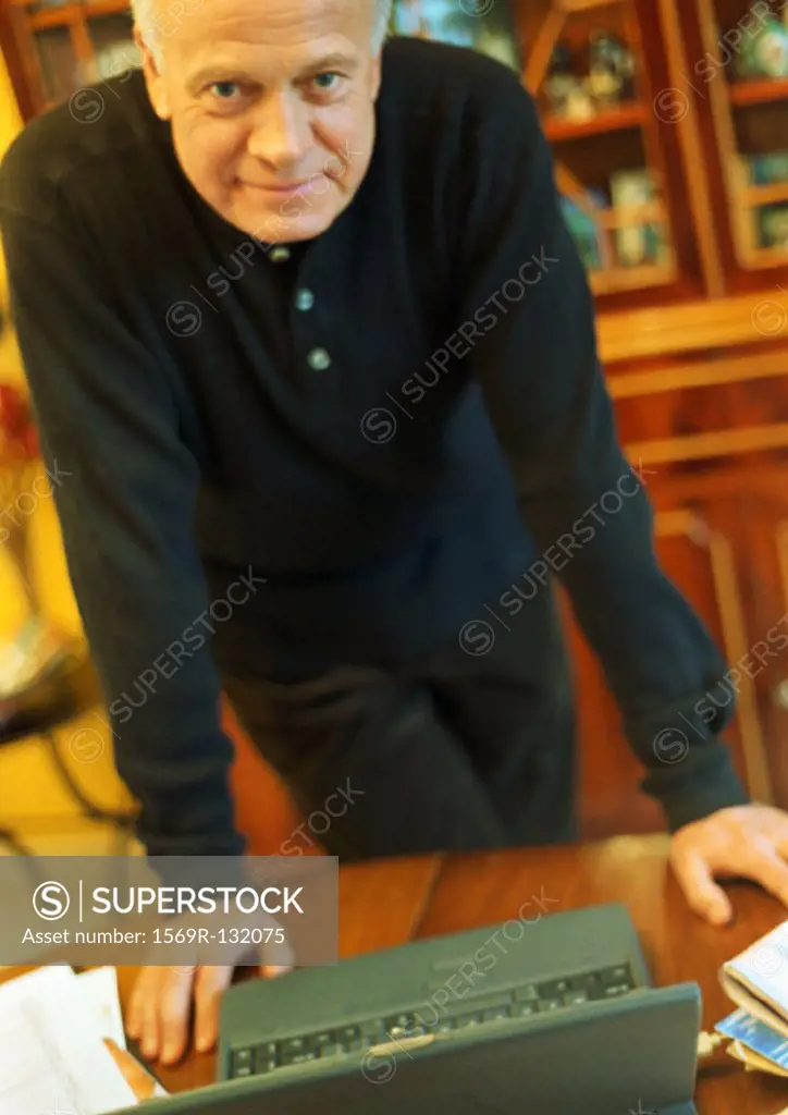 Mature man leaning on desk with laptop, looking into camera