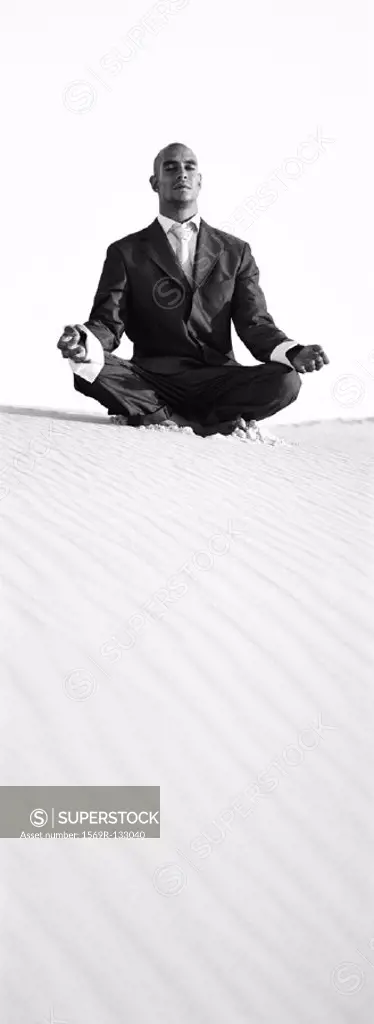 Businessman sitting in lotus position on top of dune, b&w, vertical