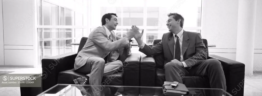 Businessmen sitting in arm chairs giving each other high five in office, b&w, panoramic view