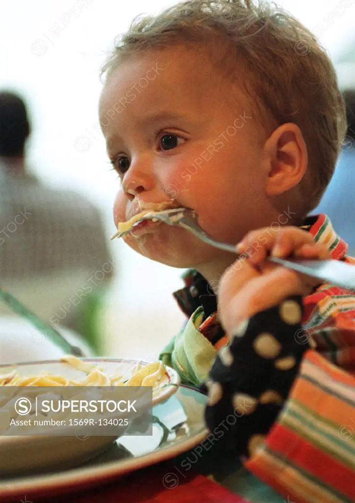 Baby, fork in mouth sideways and upside down, with bowl of pasta, head and shoulders, portrait