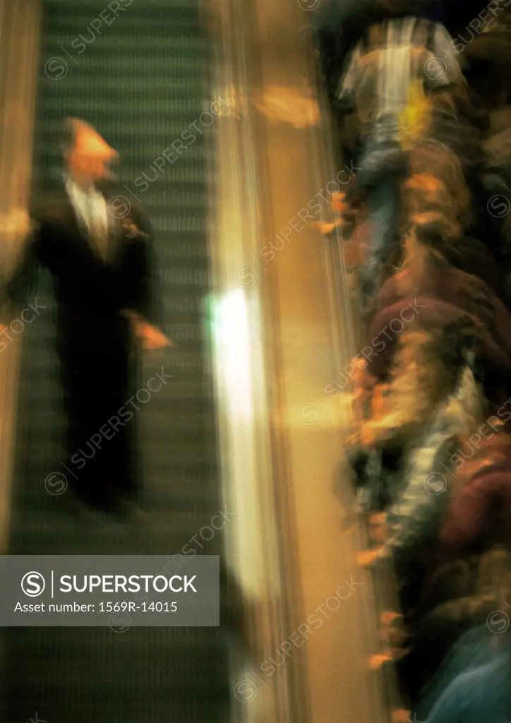 Crowd of people going up an escalator, one man coming down, blurred