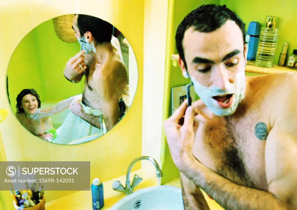 Man shaving in front of mirror, woman in reflection sitting in bathtub