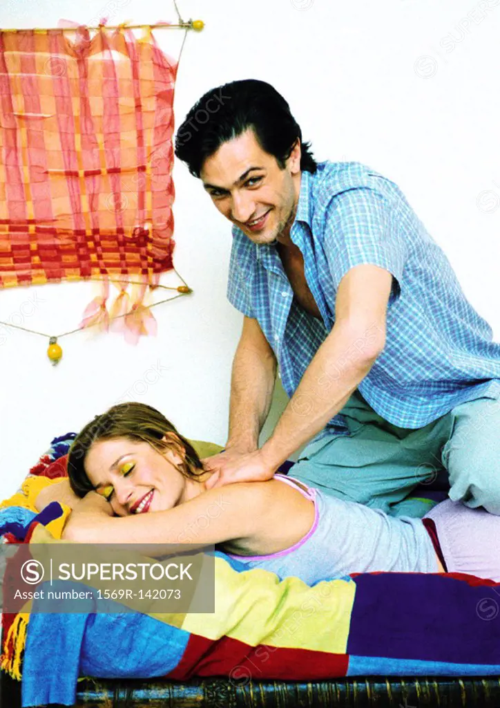 Woman lying on bed smiling, man giving her massage, looking into camera