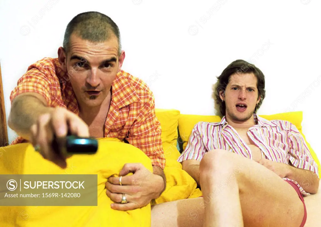 Two men sitting on sofa, one pointing remote control at camera