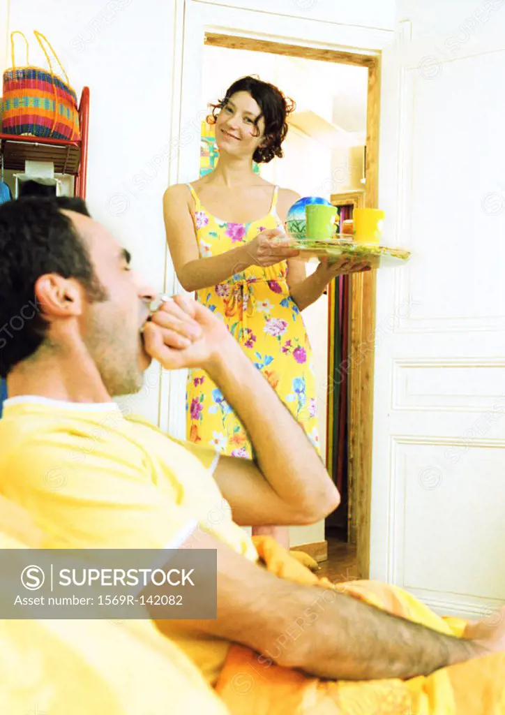 Man in bed yawning, woman carrying breakfast tray