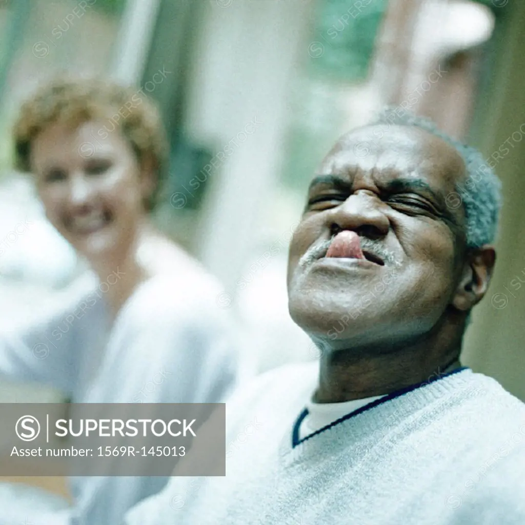 Mature man sticking out tongue, woman smiling in background