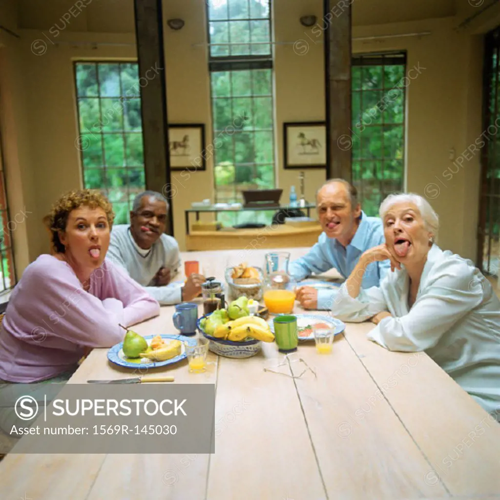 Four mature people sitting at table, sticking out tongues
