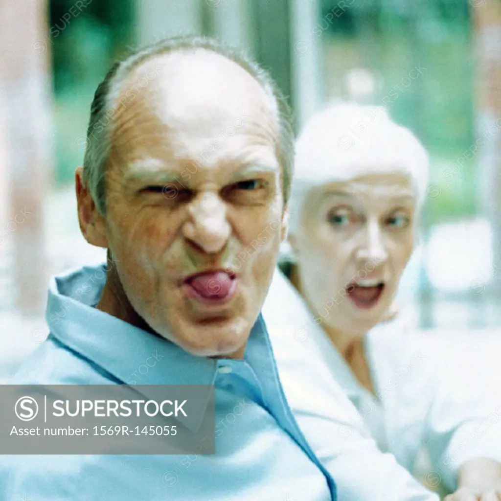 Mature man sticking out tongue, woman in background