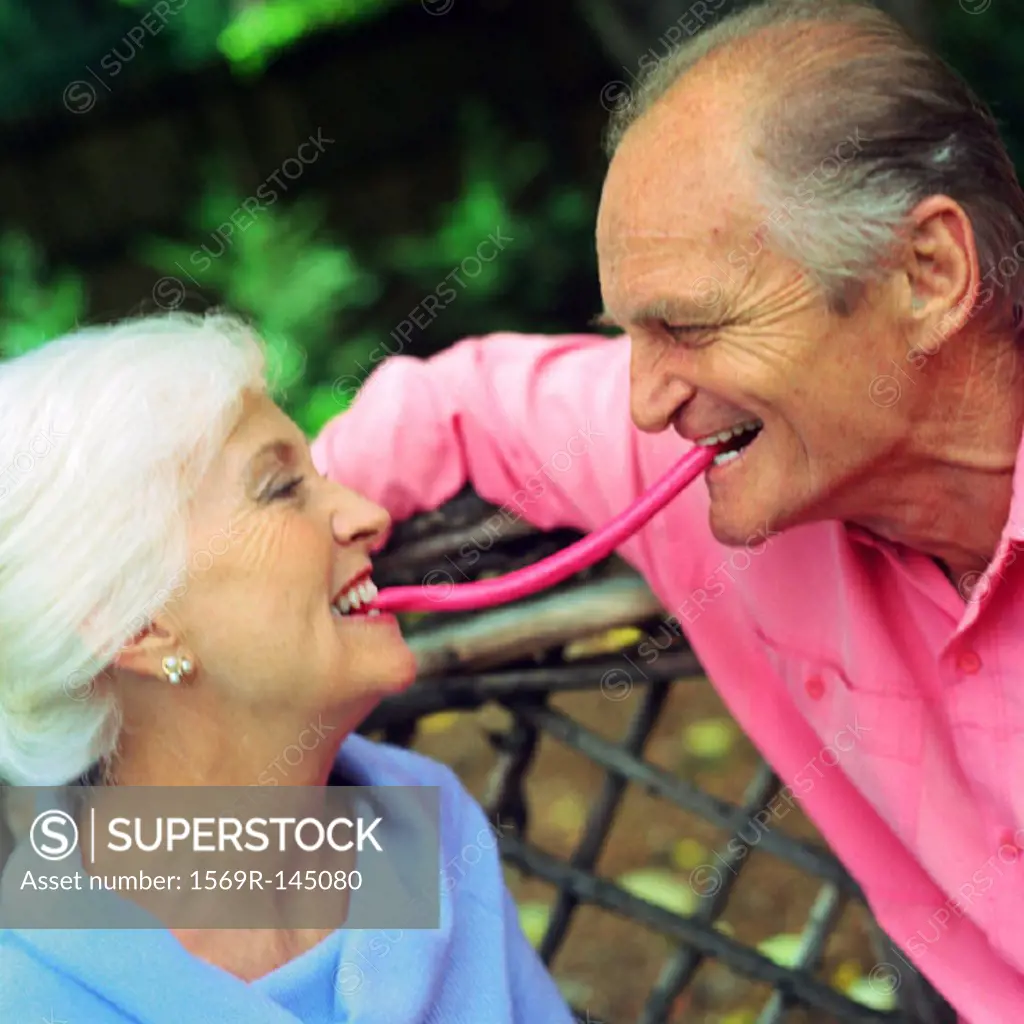 Mature couple face to face, each with one end of piece of candy in mouth between them