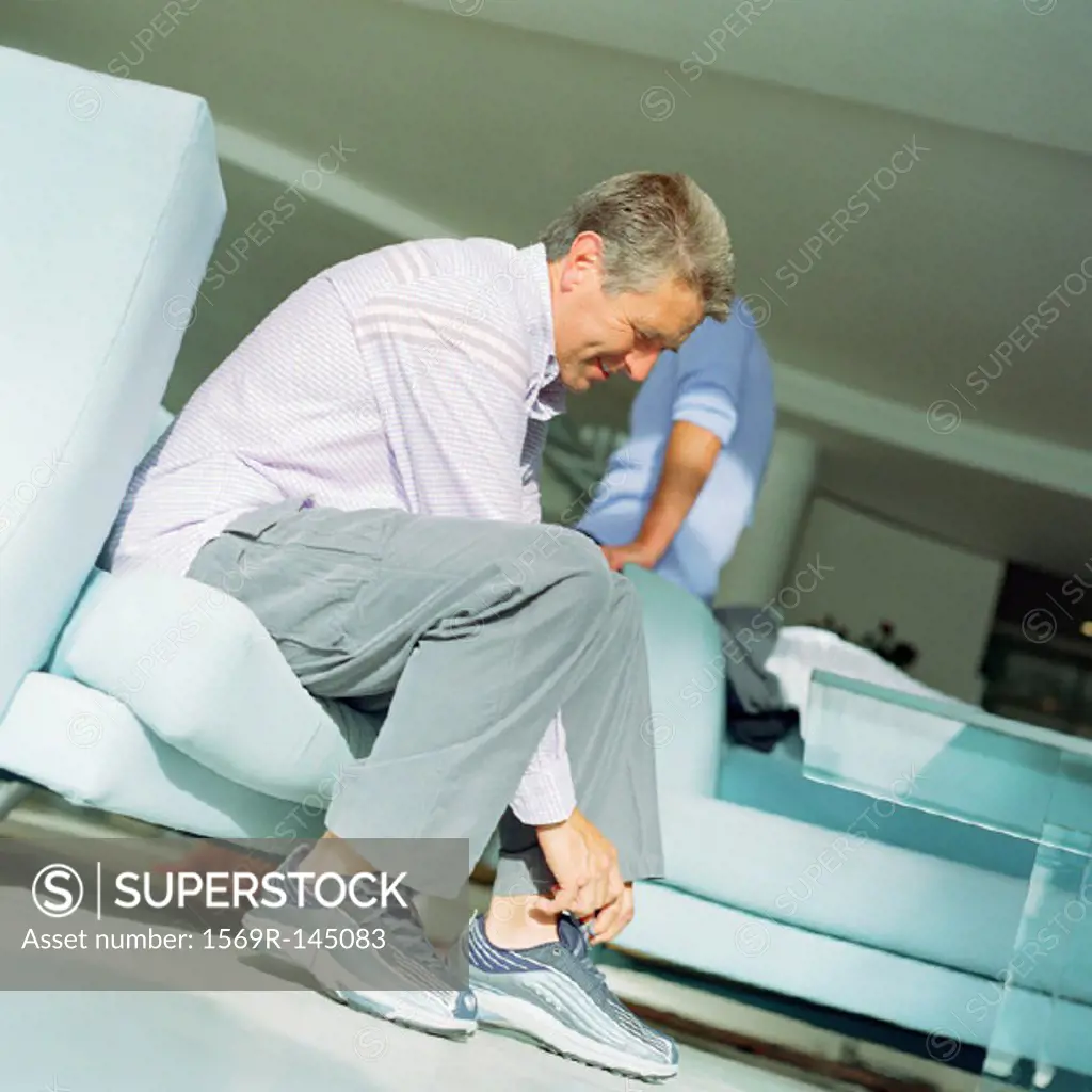 Mature man putting on shoes, second person leaning on arm of sofa, side view