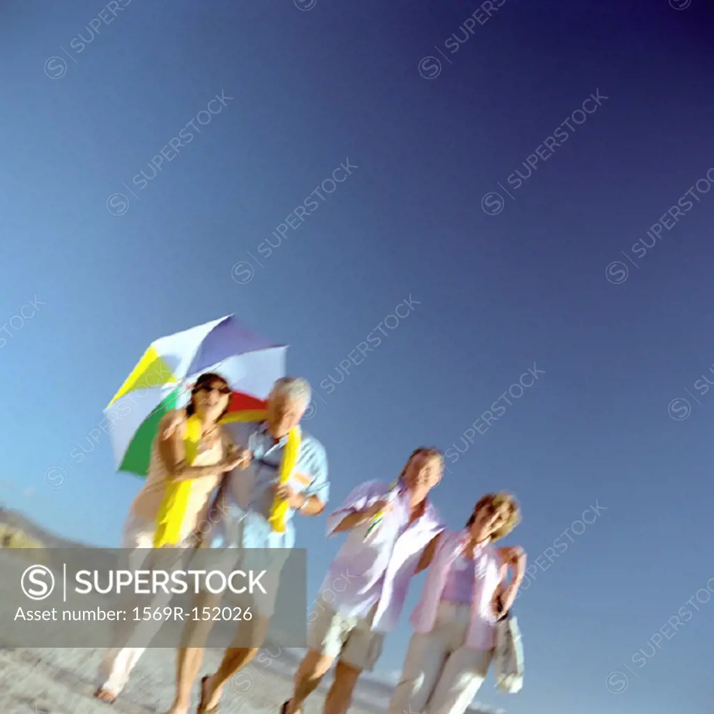 Two mature couples walking on beach, woman holding umbrella