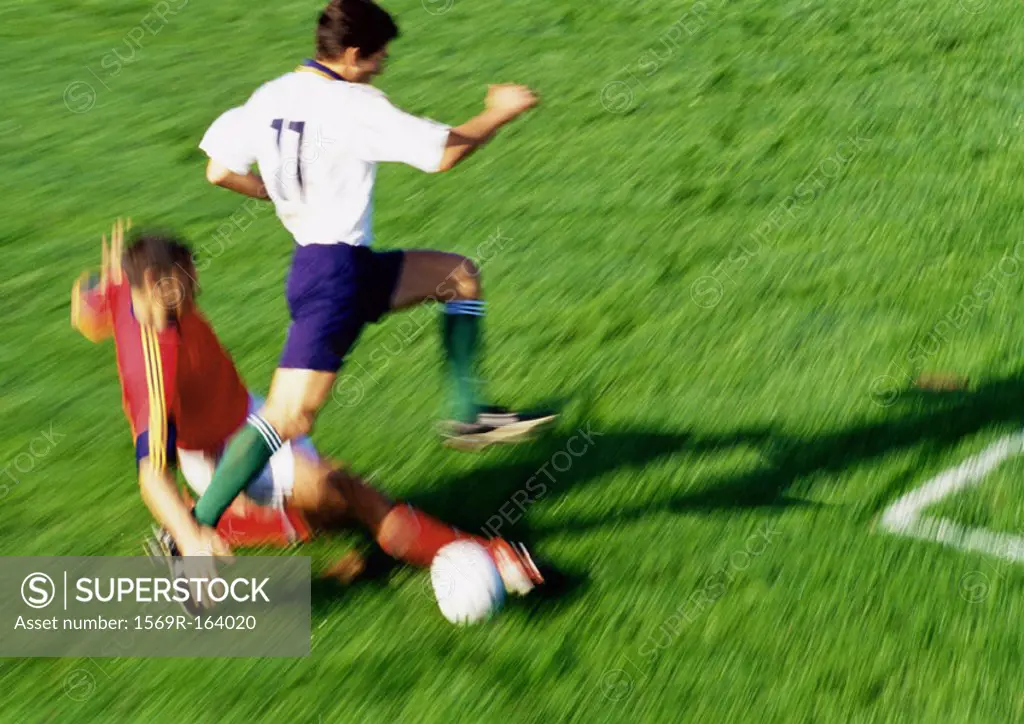 Soccer player reaching for ball, opponent jumping over leg, blurred