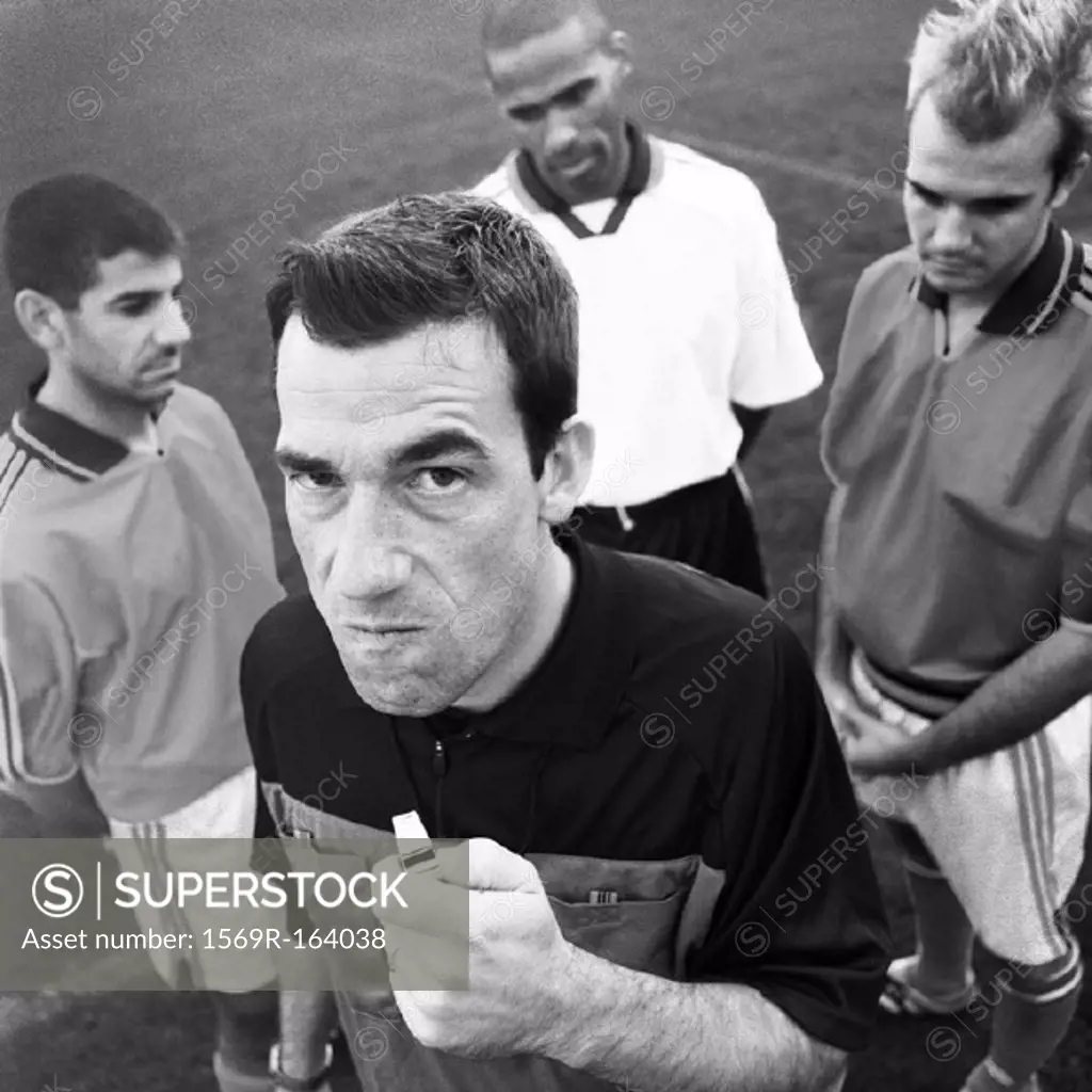 Referee holding whistle in front of three players, looking into camera, b&w