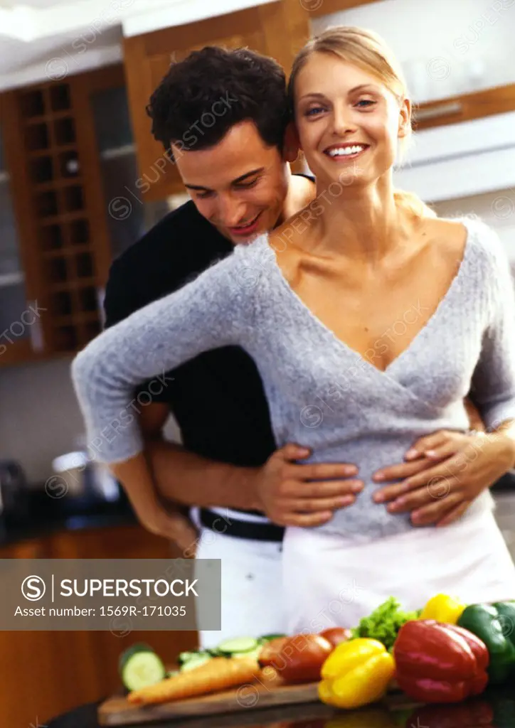 Woman standing at counter covered with vegetables, man behind woman with arms around her waist