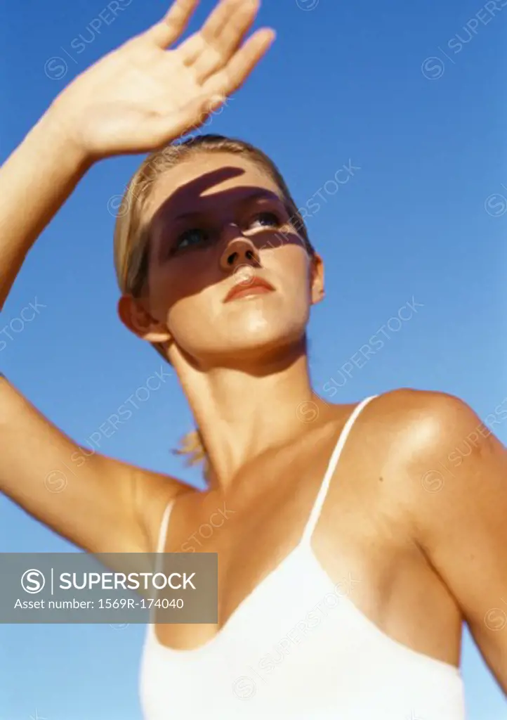 Woman protecting eyes from sun with hand, head and shoulders, low angle view