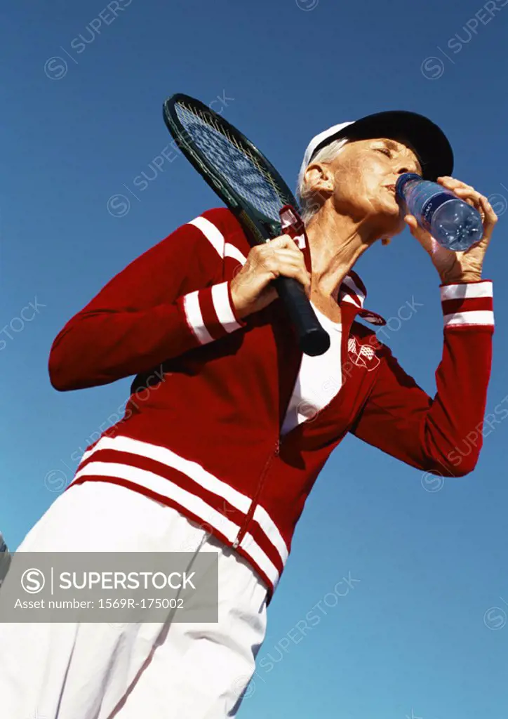 Mature woman holding tennis racket and drinking from bottle of water, low angle view