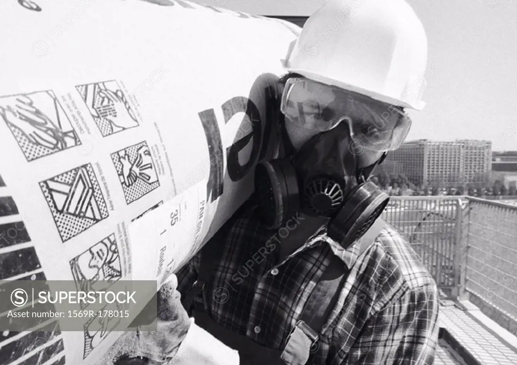 Man wearing hard hat, pollution mask and glasses, carrying roll of insulation on shoulder, close-up, b&w