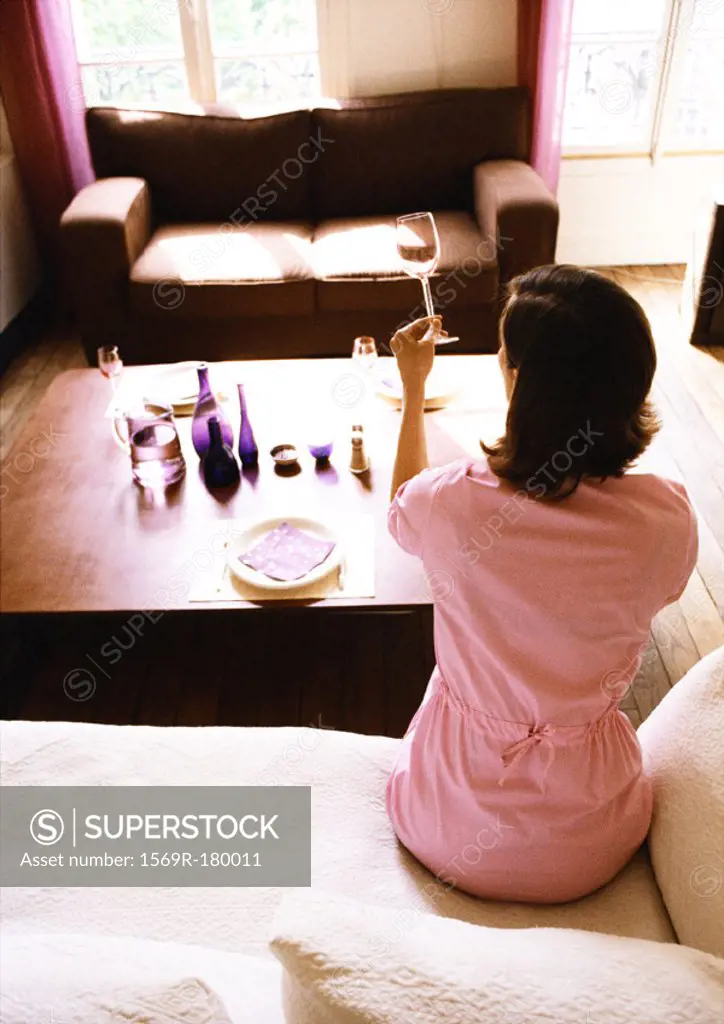 Woman sitting in living room, holding up wine glass, rear view