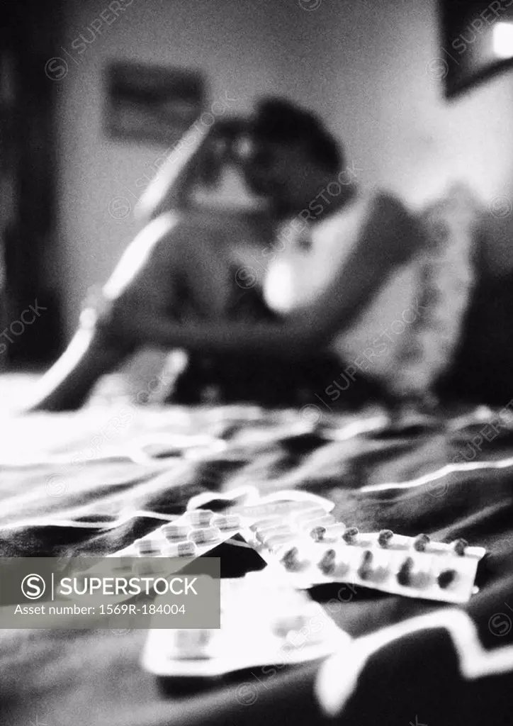 Woman sitting on bed, pills in foreground, blurred, b&w
