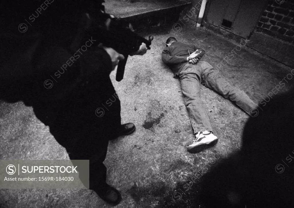Man standing and pointing sub-machine gun at man face-down on floor, b&w