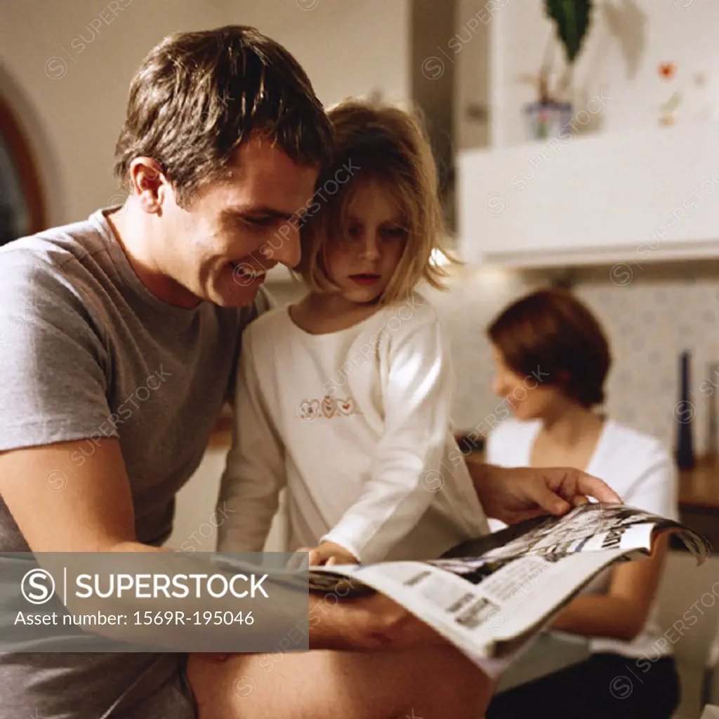 Father sitting with daughter looking at paper, woman in background