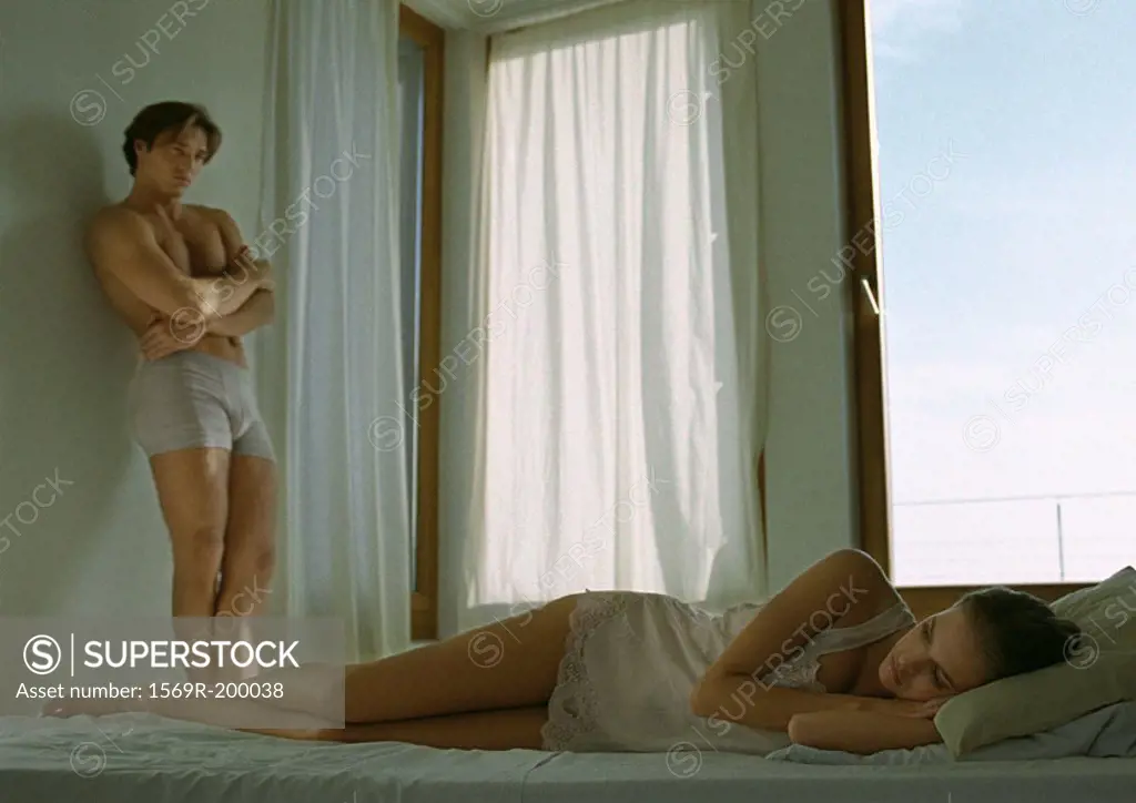 Woman lying in bed, man standing against wall looking at her