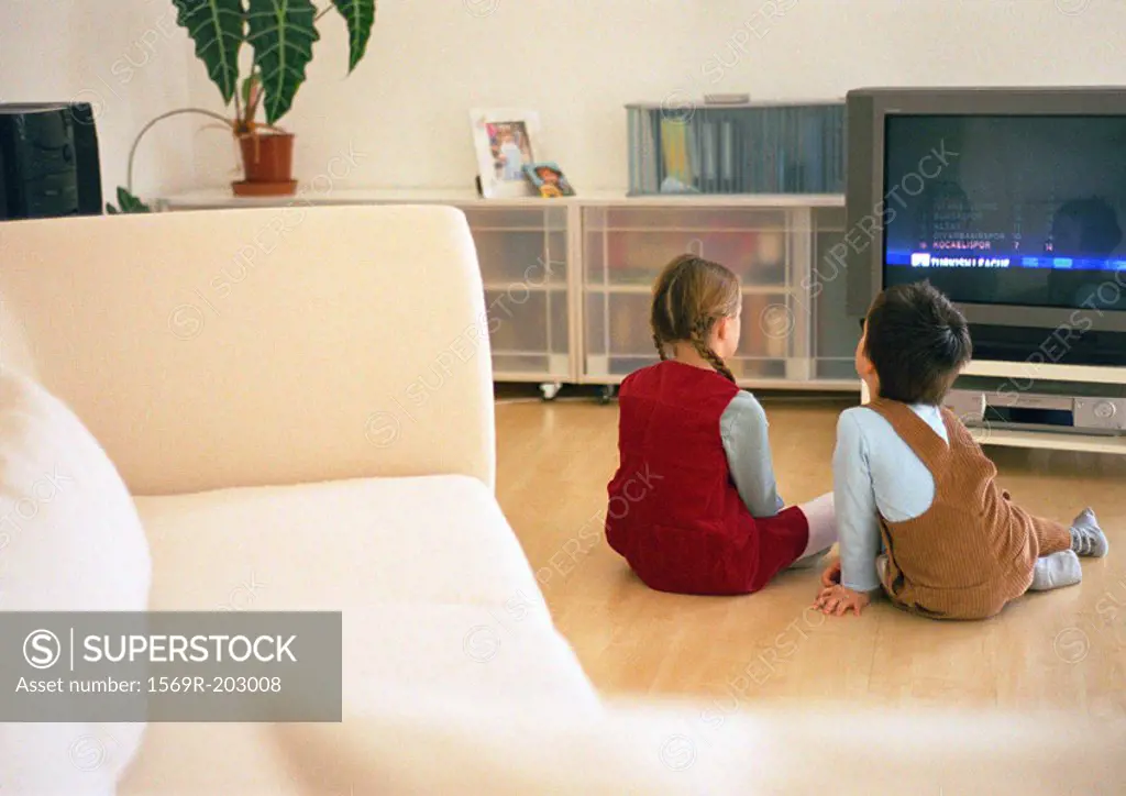 Lttle boy and girl sitting together on parquet floor looking at tv, rear view