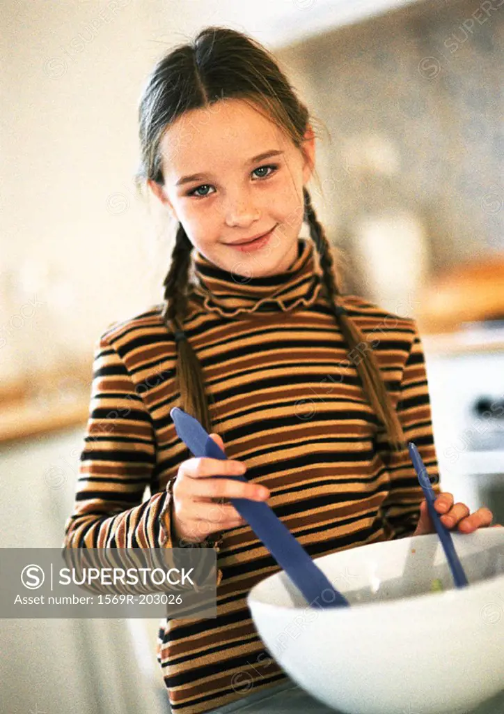 Young girl waist up, standing behind white mixing bowl, holding utensils in bowl, smiling, in kitchen setting, tilt