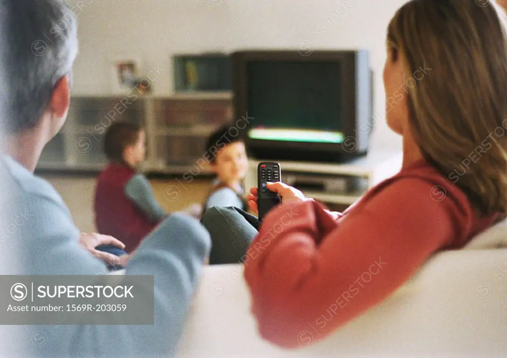 Adult man and woman sitting on sofa, rear view, blurred in foreground, children near tv in background