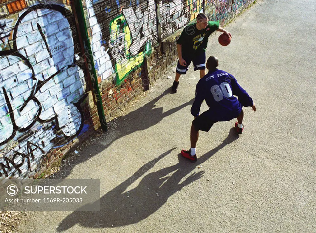 Two men playing basketball in front of graffitied wall in urban playground