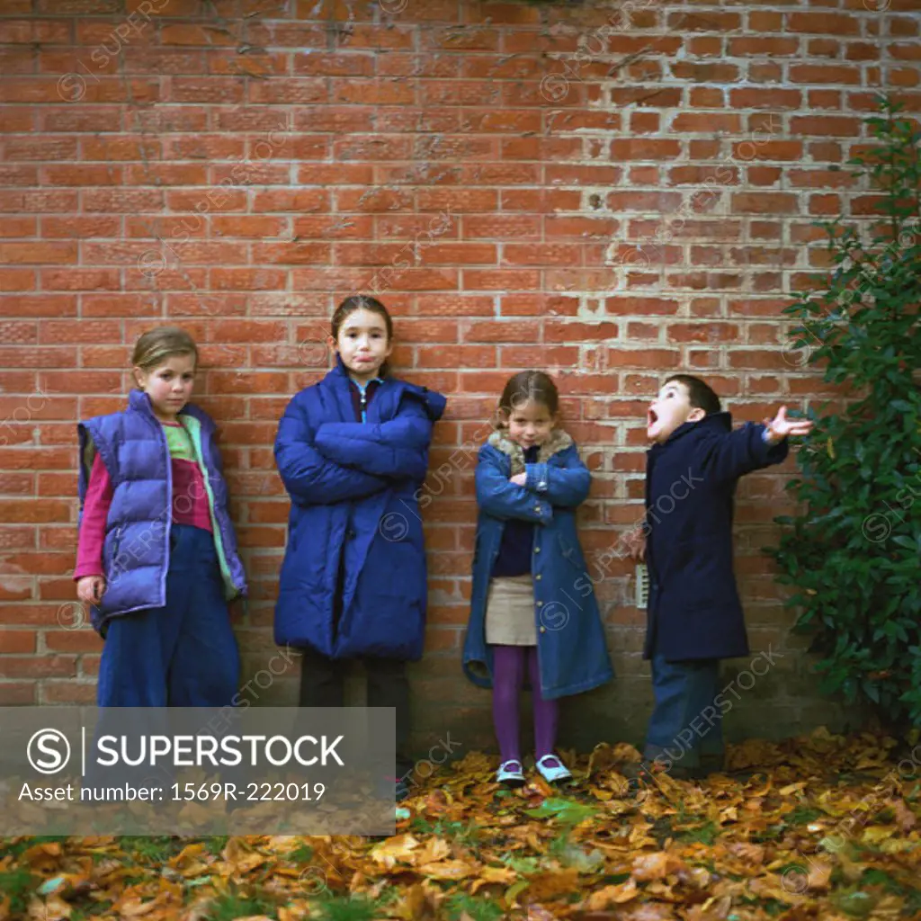 Four children wearing coats in front of brick wall, full length