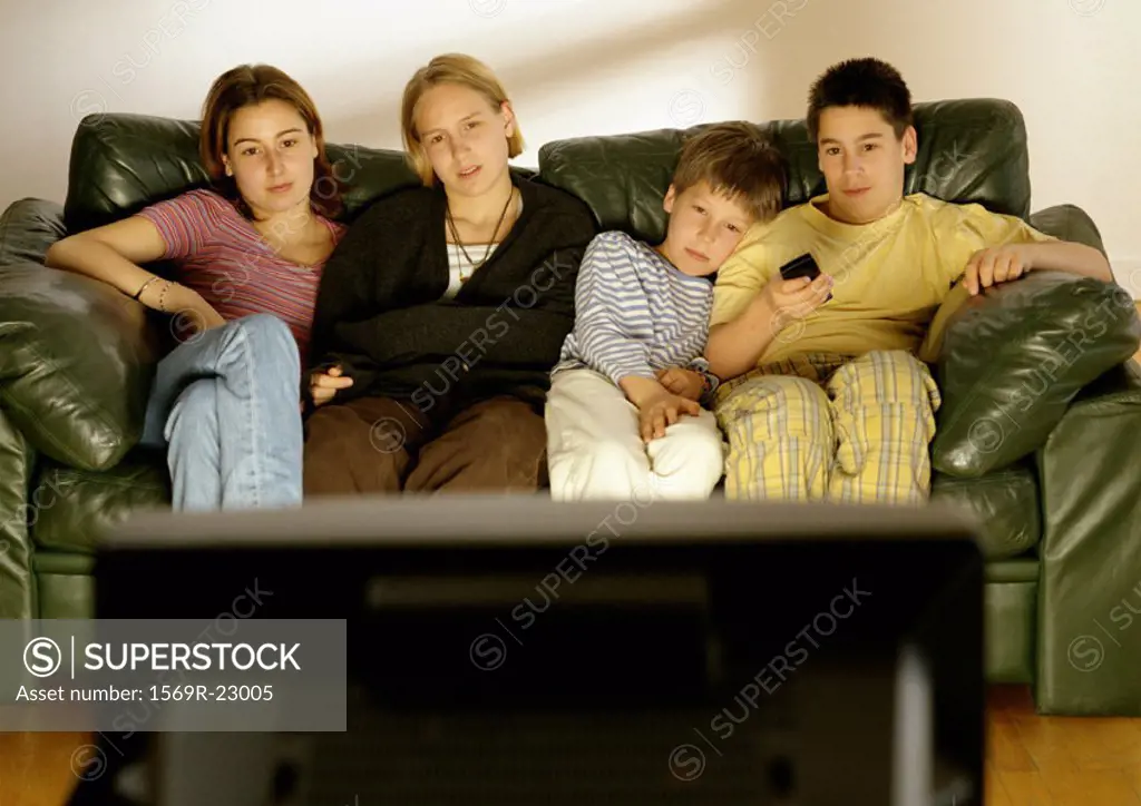 Focus on young adults and children sitting on couch in background, rear view of television in foreground, blurred