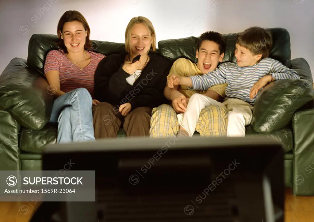 Three young people and one child sitting on green couch together, laughing, blurred rear view of tv in foreground