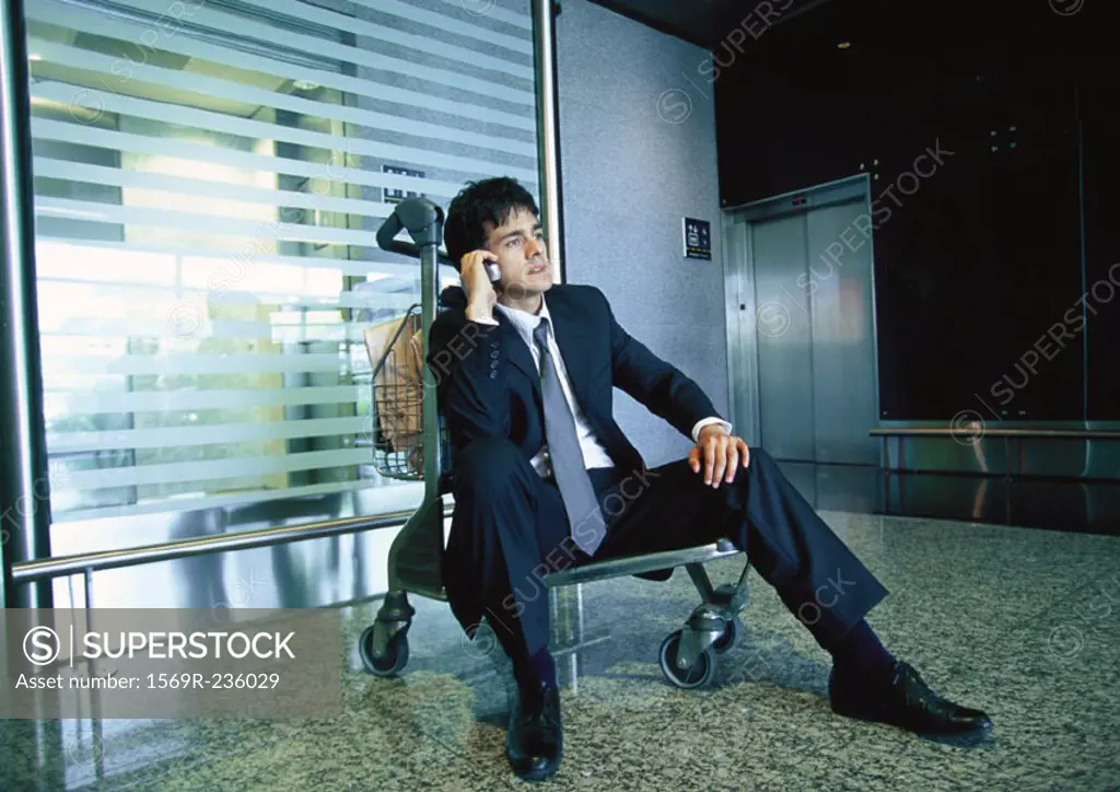 Businessman sitting on luggage cart talking on cell phone