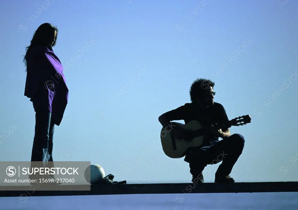 Woman standing, man squatting, playing guitar on bench