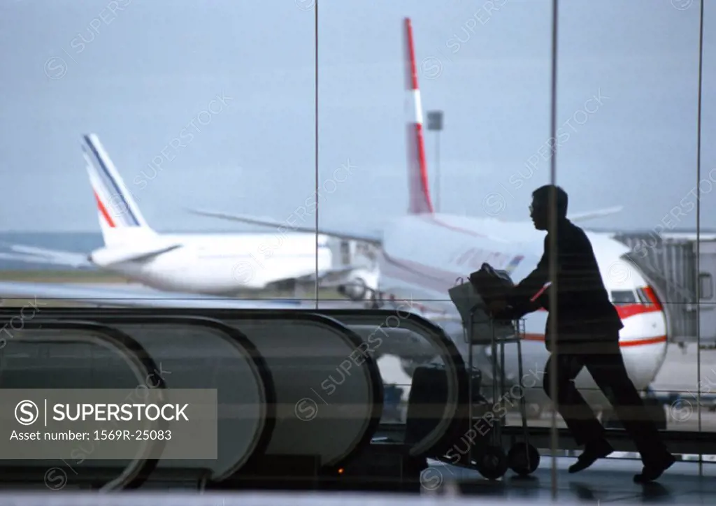 Silhouette of man pushing luggage cart, view through glass window, airplanes in background