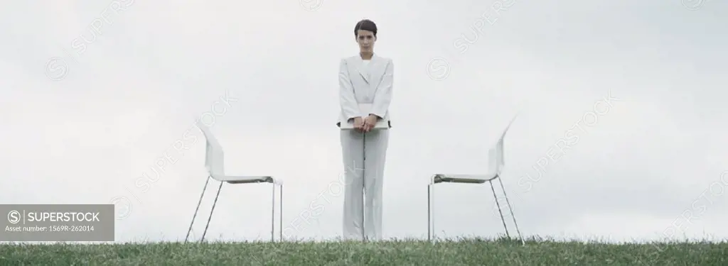 Woman standing outdoors on grass between two chairs in front of overcast sky
