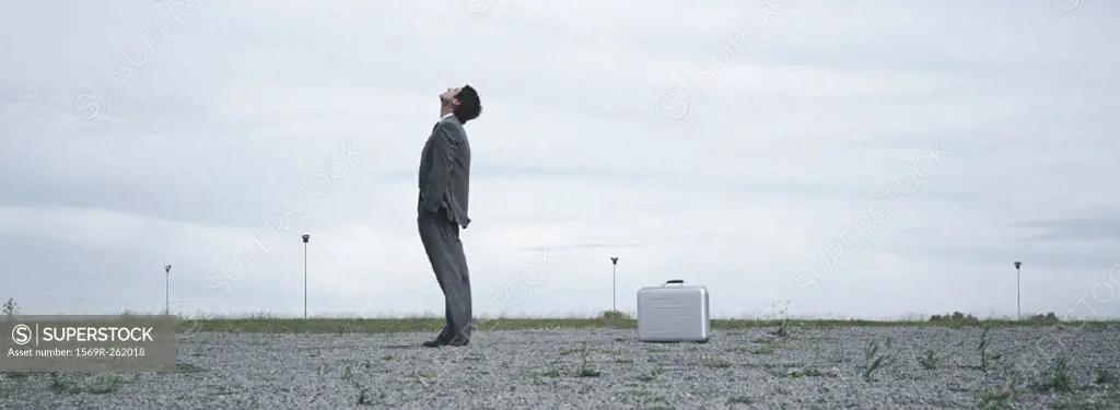 Man standing in abandoned lot looking up at sky, metallic briefcase on ground behind him