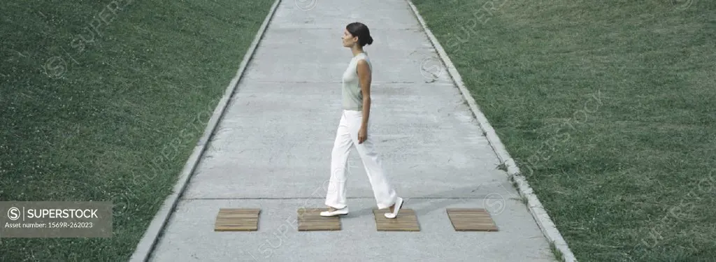 Woman stepping on wood squares across concrete path, full length