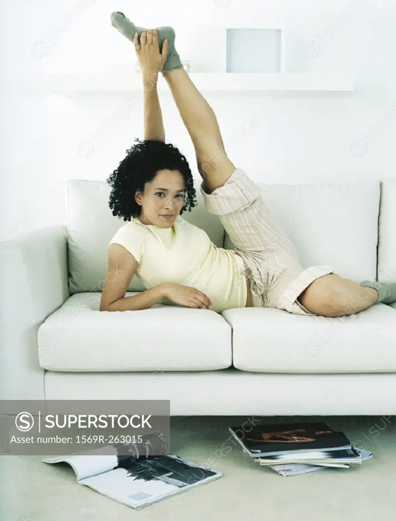 Woman lying on sofa leaning on elbow and stretching leg, looking at camera