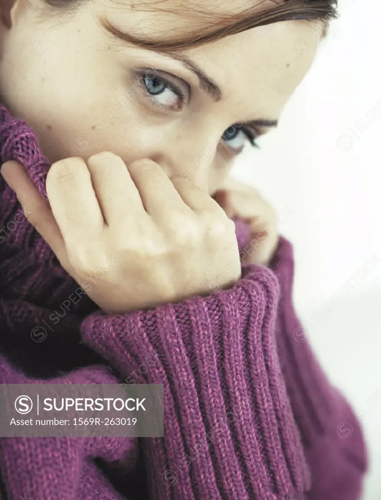 Woman covering part of face with neck of sweater, looking at camera, close-up