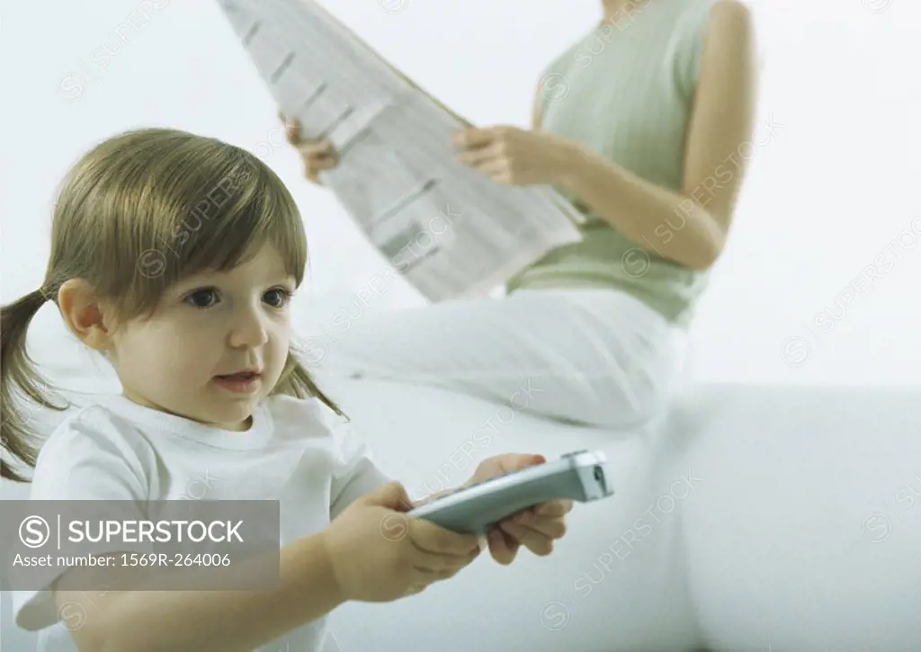 Little girl pointing remote control, woman sitting on back of sofa holding newspaper in background