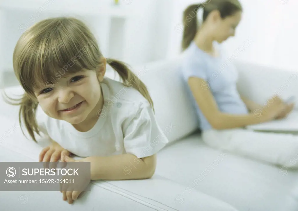 Little girl leaning on sofa arm making faces at camera, young woman looking down in background