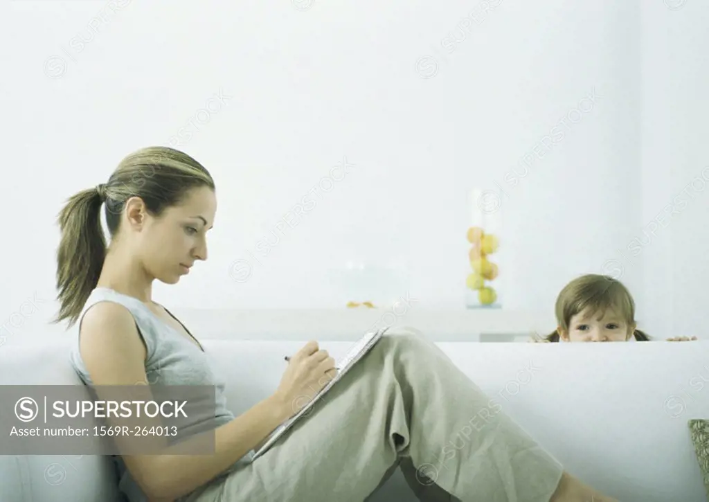 Young woman sitting on sofa writing on notepad, girl peeking over edge of sofa