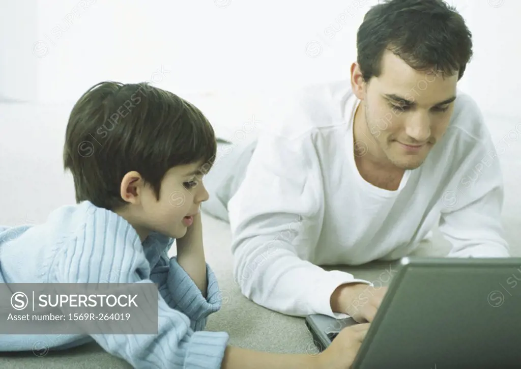 Man and little boy lying on floor on stomachs, working on laptop