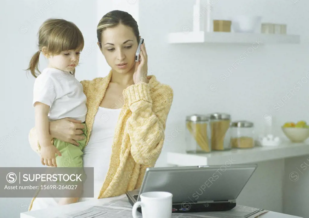 Young woman holding little girl with one arm, holding cell phone to ear with other hand, looking down at laptop