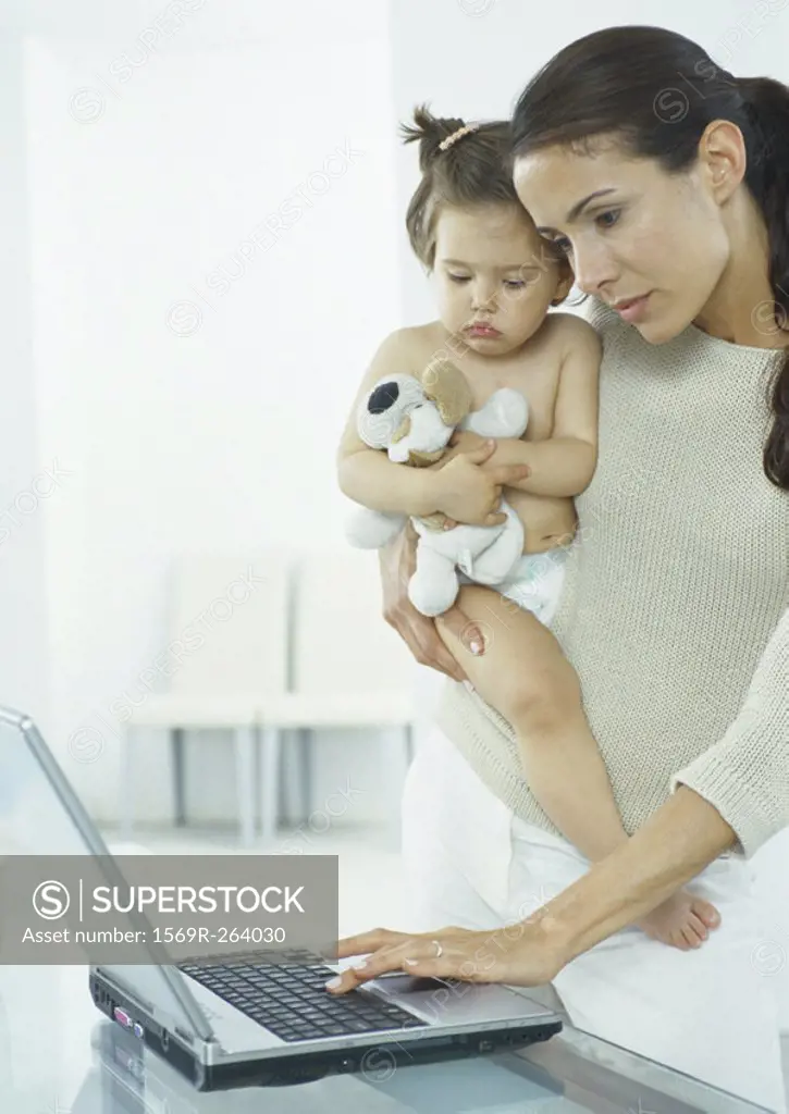 Woman holding little girl in one arm, typing on laptop with other hand