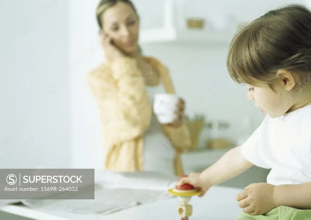 Little girl playing with toy, woman holding mug and talking on phone in background