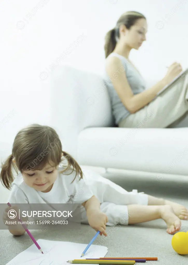 Little girl lying on floor coloring, young woman sitting behind her on couch writing with knees up