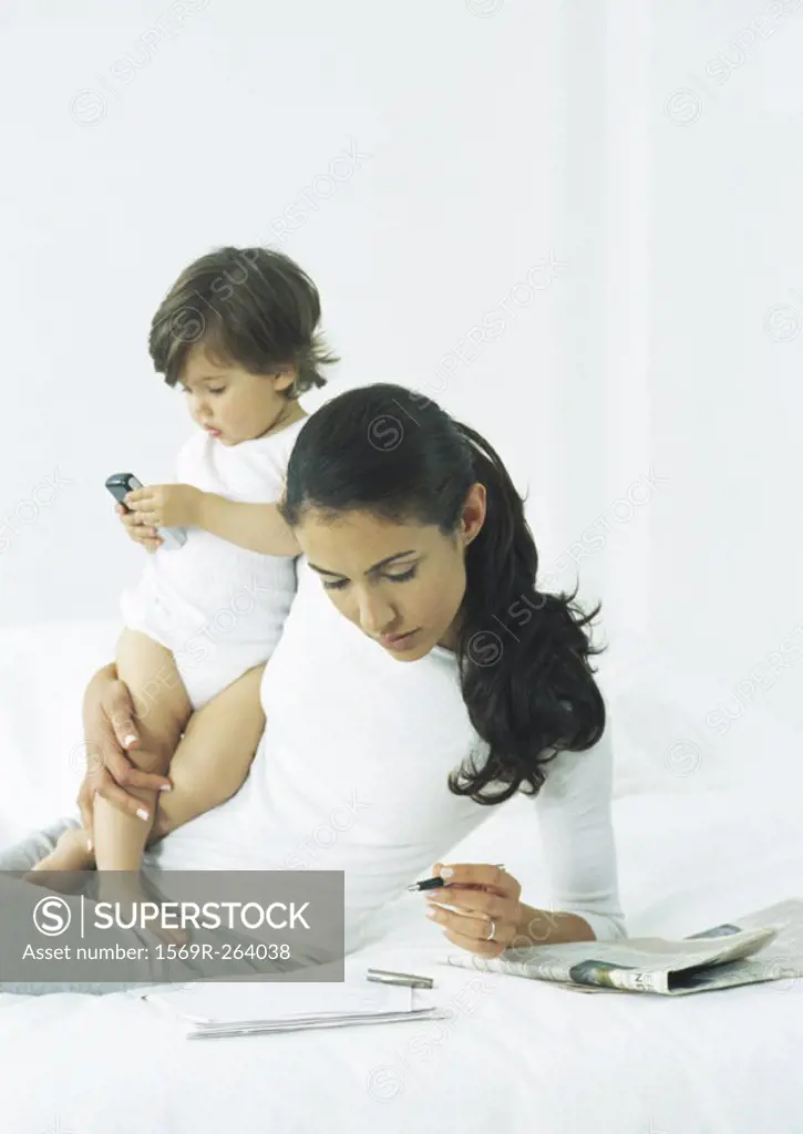 Young woman propped up on elbow holding pen with one hand, holding little girl with other arm